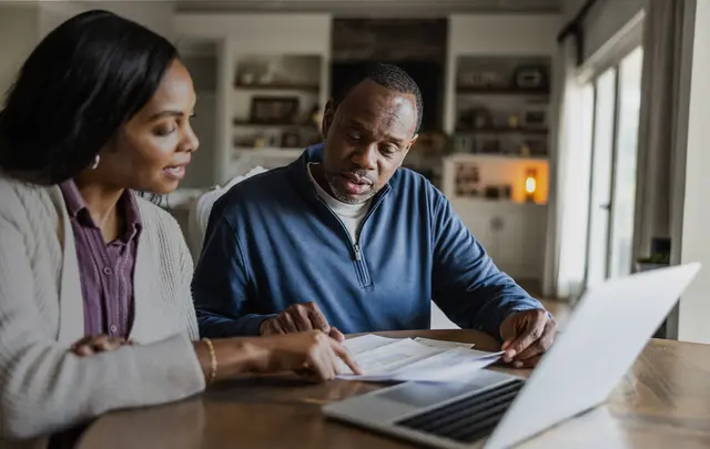 Two people sitting at a table reviewing printed documents together, with an open laptop nearby, in a bright home or office setting.