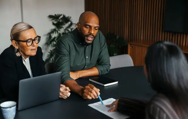 Three people seated at a meeting table reviewing a document together, with one person pointing at the page while another takes notes on a laptop.