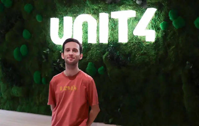 Man standing in front of a green moss wall with a bright illuminated “UNIT4” sign, wearing a red Levi’s T‑shirt in an indoor office setting.