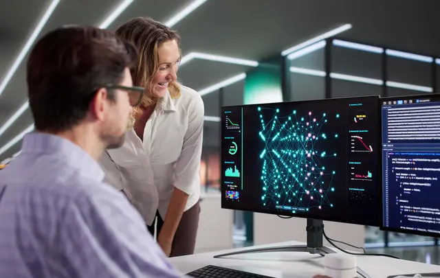 Two professionals collaborating at a desk, reviewing data visualizations and code on a widescreen monitor displaying a network graph and analytics dashboards in a modern office environment.