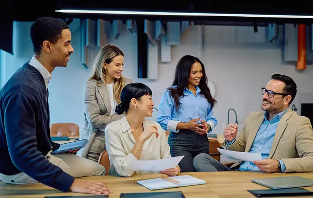 A group of professionals collaborating in a modern office setting, reviewing documents and discussing insights around a table, with laptops and materials visible, highlighting teamwork, planning, and decision-making.