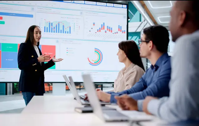 Professional presenting data on a large digital dashboard with charts and graphs to a group seated at a conference table, using laptops in a modern office setting.