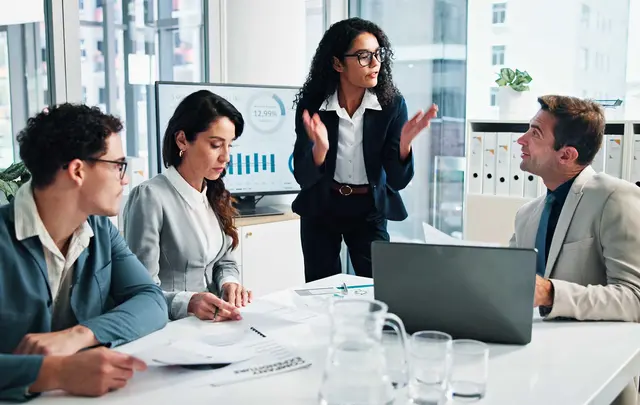 Four professionals in a modern office meeting room sit around a table reviewing printed documents and a laptop, while one person stands and gestures during a discussion. A screen behind shows charts and data, with binders and glassware visible on the table.