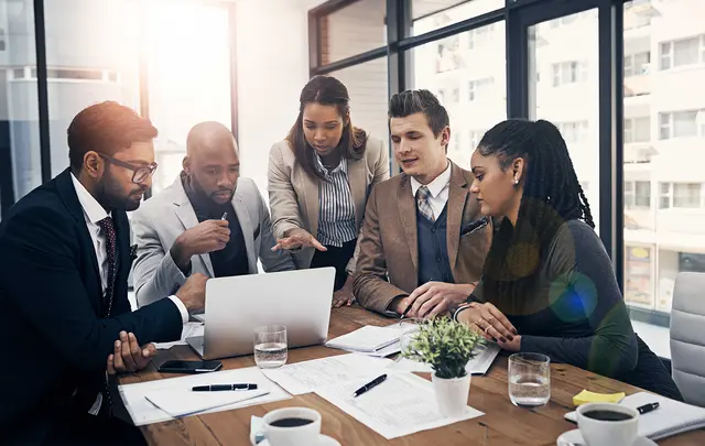 Diverse team collaborating in a modern office around a laptop, reviewing documents and financial data together, with notebooks, coffee and a bright windowed workspace in the background.