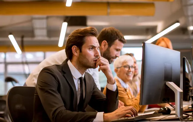 Professional office setting with a group of employees working at desktop computers; a person in the foreground focuses on a screen while colleagues collaborate behind, in a modern workspace with warm lighting.