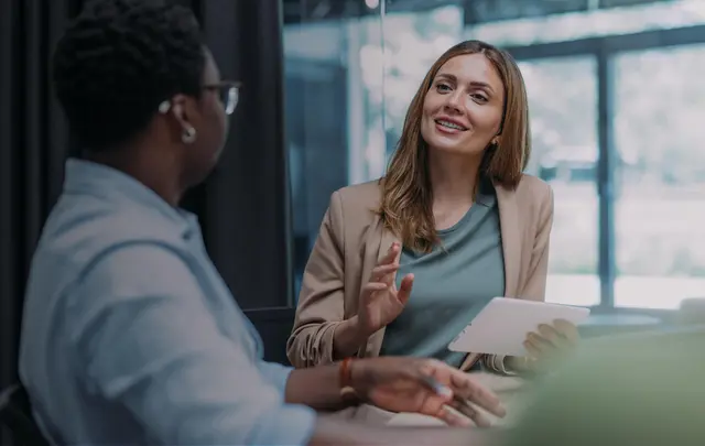 Two professionals in a modern office environment engaged in a discussion, with one person holding a tablet and explaining information while the other listens attentively. 