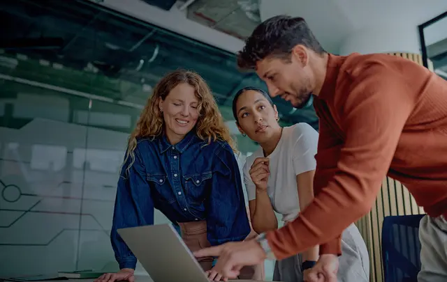 Three colleagues collaborating around a laptop in a modern office, reviewing information together in a bright workspace with glass walls and contemporary furnishings.