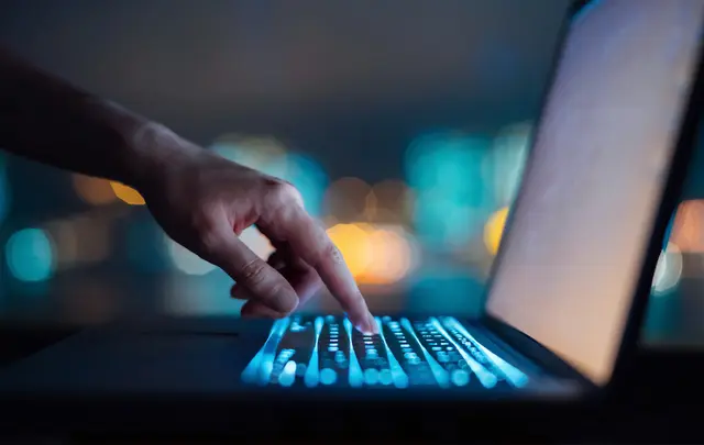 Close-up of a hand typing on a laptop keyboard with glowing blue keys in a low-light setting, representing data analysis or AI-driven work.