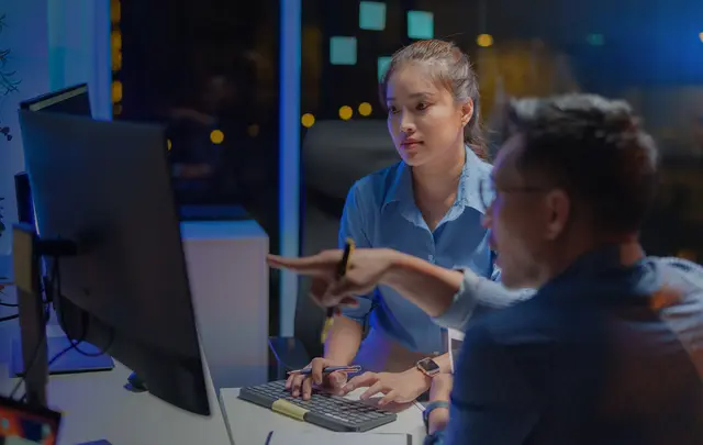 Two colleagues working together at a computer in an office at night, with one pointing at the screen while the other types on a keyboard.