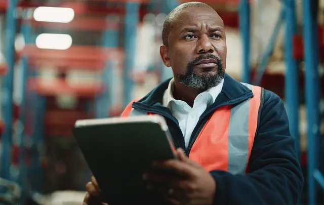 Worker wearing a high-visibility orange safety vest holding a tablet in a warehouse or industrial setting, reviewing data or inventory.