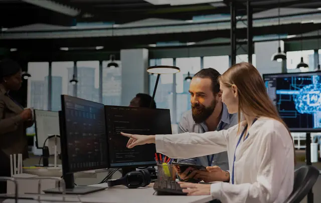 Two professionals collaborating at desks in a modern office, reviewing data and code on multiple monitors, with the foreground screen displaying dashboards and analytics, and a cityscape visible through large windows in the background.