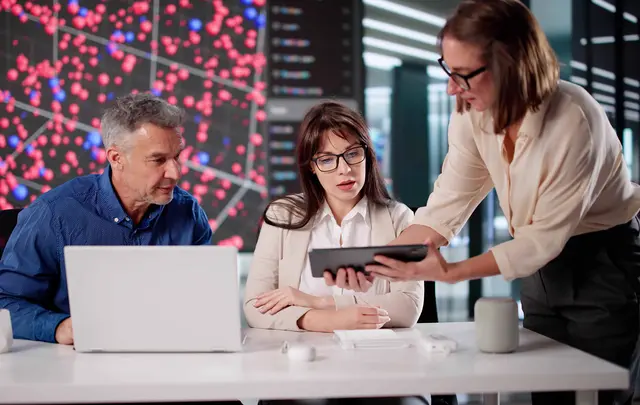 Three professionals collaborate at a desk in a modern office, reviewing information on a tablet and laptop.