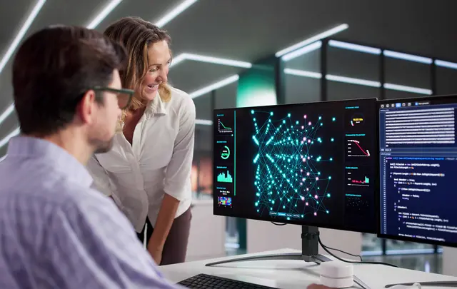 Two professionals review workforce and organizational data on dual computer monitors in a modern office.
