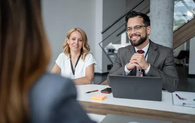 Two professionals sit across a table in a modern office meeting space, facing a person in the foreground.