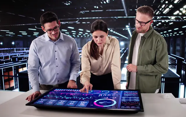 Three professionals collaborate around a large interactive touchscreen displaying procurement and business analytics dashboards with charts, graphs, and performance metrics.