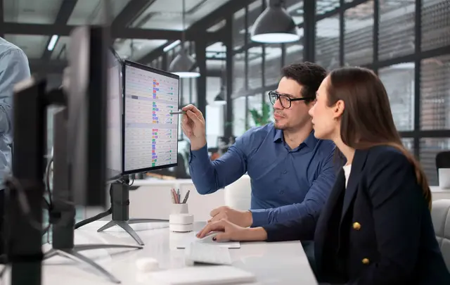 Two professionals sit at a desk in a modern office, reviewing financial or operational data displayed on a computer monitor.