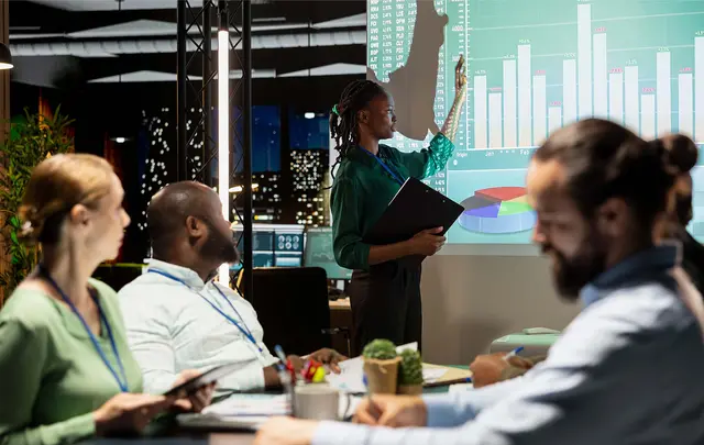 Person presenting workforce analytics and performance dashboards on a large screen during a business meeting, while colleagues review data and reports around a conference table.