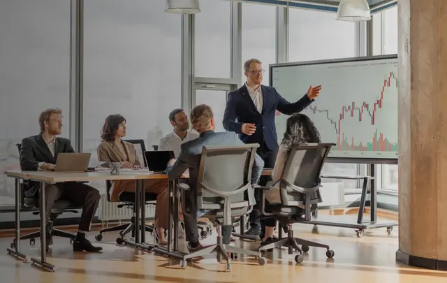 Business professionals gathered in a meeting room while a presenter discusses financial performance data displayed on a large screen, illustrating strategic planning, AI-driven finance insights, and decision-making.