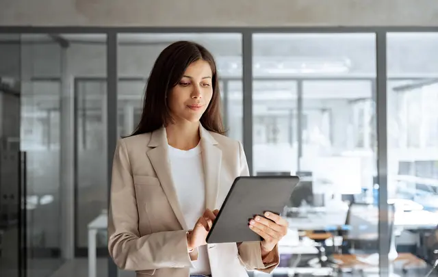 A professional standing in a modern office with glass walls uses a tablet device.