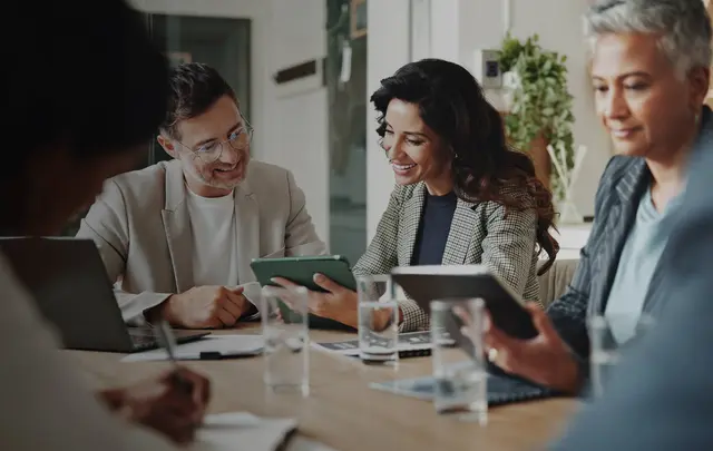 A group of professionals seated around a conference table collaborate using digital devices during a meeting.