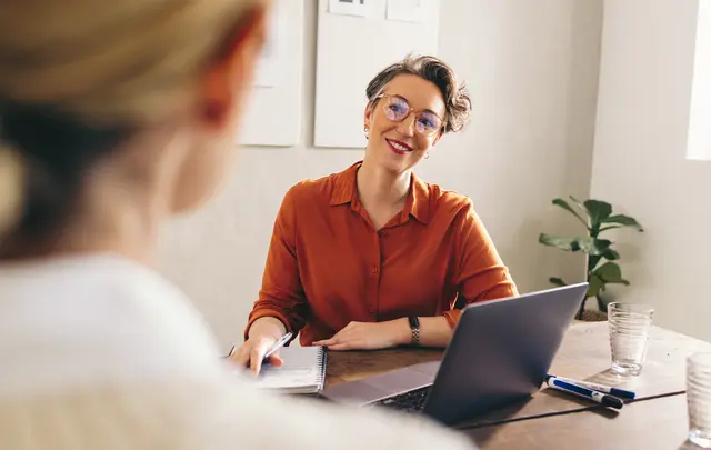 Person wearing an orange shirt and glasses sits at a desk with an open laptop, taking notes during a one-to-one workplace meeting. 
