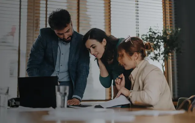 Three colleagues gathered around a laptop and printed documents in a modern office, reviewing procurement data and discussing business decisions during a collaborative planning session.