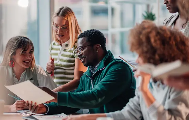 A group of colleagues collaborating around a table in a modern office, reviewing printed documents and discussing ideas during a team meeting.