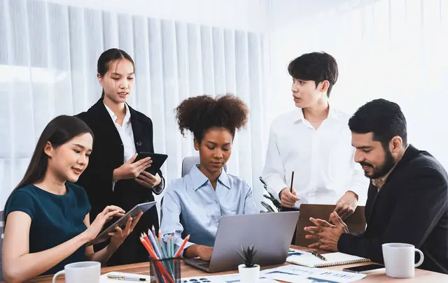 Team collaborating around a laptop during a procurement planning meeting, with participants reviewing documents, using tablets, and discussing workflow processes in a modern office environment.