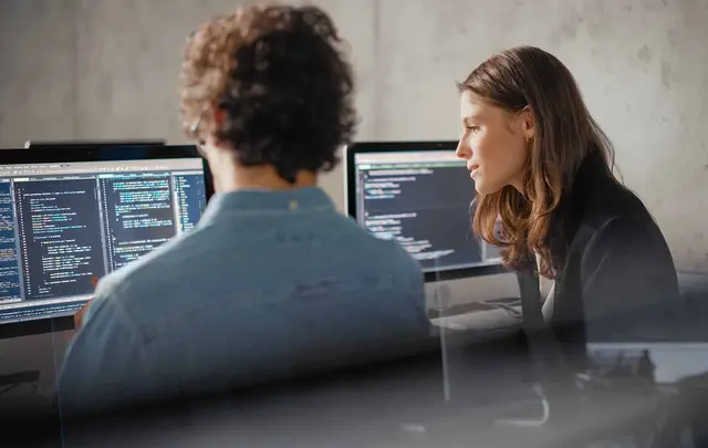 Two professionals collaborating at a workstation, reviewing software code displayed on multiple computer monitors in a modern office environment.