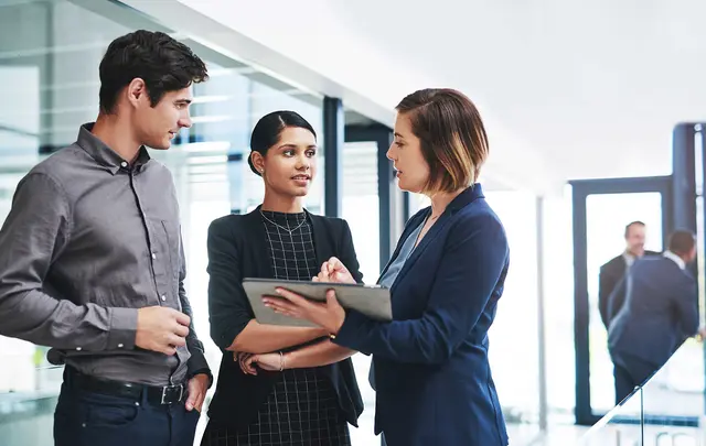 Three business professionals standing in a modern office discussing financial planning and decision-making while reviewing information on a tablet.