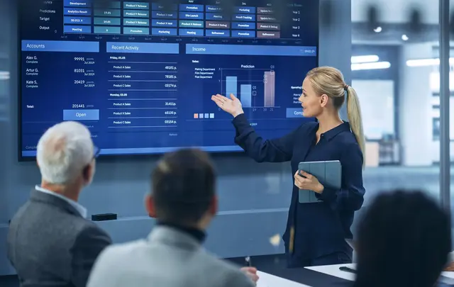 Presenter explaining multi-entity finance performance data on a digital analytics dashboard with revenue charts, forecasts, and financial reporting insights during a business meeting.
