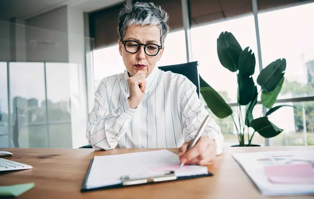 Professional seated at a desk in a modern office, reviewing documents and making notes on a clipboard.