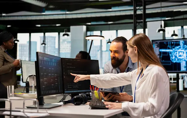 Two professionals collaborate at a workstation in a modern office, reviewing data displayed on multiple computer monitors.