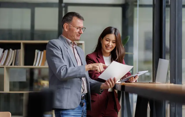 Two professionals standing in a modern office workspace reviewing printed documents together beside a workstation.