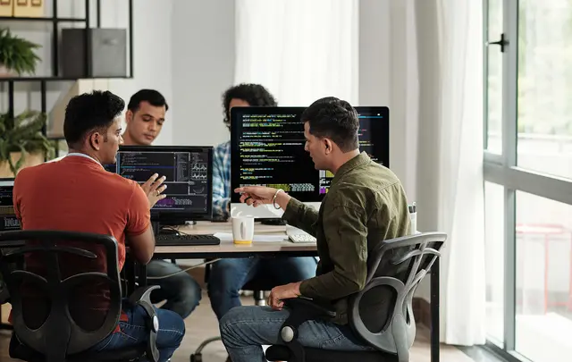 Team members collaborating in a modern office, reviewing code and data dashboards displayed on multiple computer monitors during a software development or technology planning session.