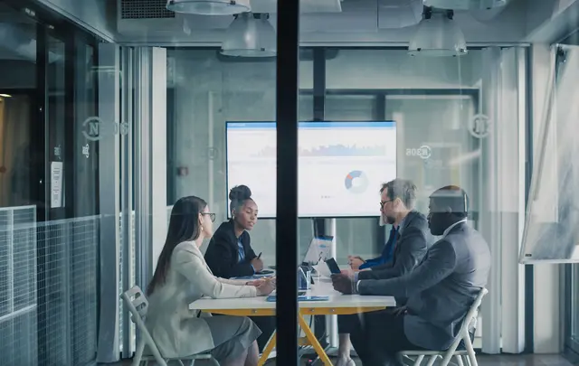 Professionals meeting in a modern glass-walled conference room, reviewing business performance dashboards and charts displayed on screens during a collaborative planning session.
