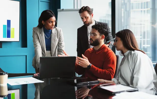 Four professionals collaborate around a laptop in a modern office meeting room.
