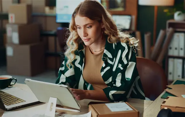 Person working at a desk in a modern office, using a tablet beside a laptop and shipping boxes, reviewing information and managing supplier or procurement-related tasks.