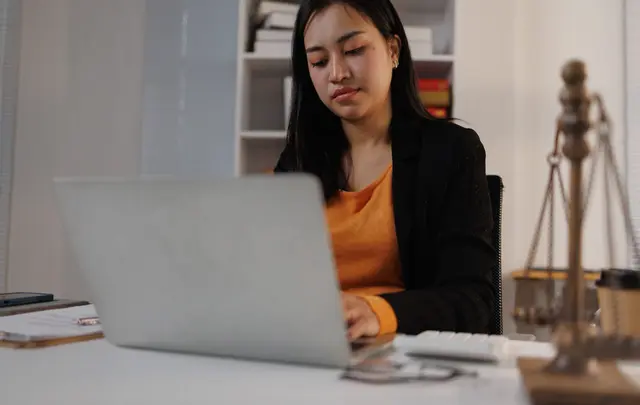Person working on a laptop at a desk in an office setting, with a scales of justice ornament in the foreground, bookshelves in the background, and office accessories on the desk.