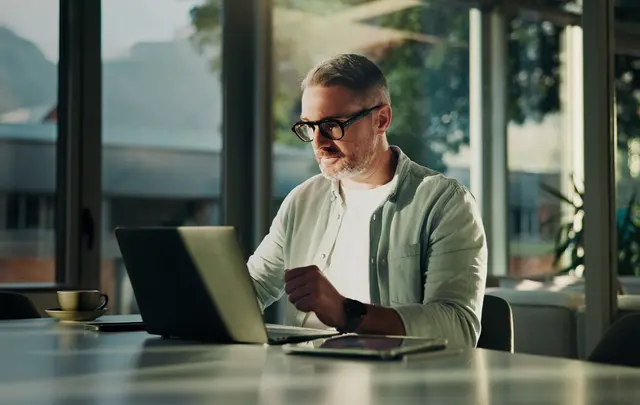 Professional working on a laptop at a desk in a modern office with large windows and natural light. A tablet and coffee cup sit on the table, while the individual reviews information on the screen in a bright, contemporary workspace.
