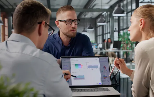 Three professionals review workforce planning and people cost analytics on a laptop dashboard during a meeting in a modern office.