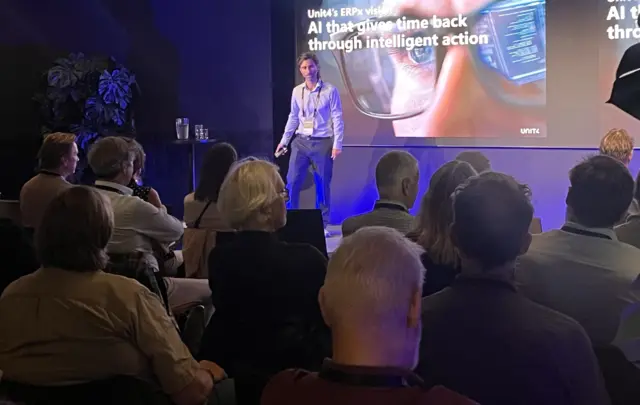 Presenter speaking on stage at a business conference while attendees watch from the audience.