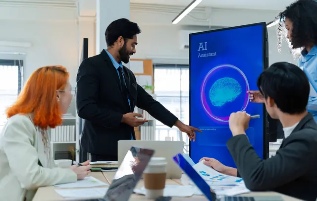 A group of professionals gathers around a conference table in a modern office while one presenter demonstrates an AI assistant interface on a large digital screen.