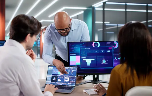 Three professionals collaborate in a modern office while reviewing business performance dashboards displayed on a laptop and external monitor.