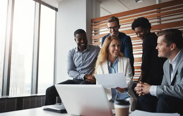 A group of business professionals gathered in a modern office meeting area, reviewing documents and collaborating around a laptop during a planning or strategy discussion.