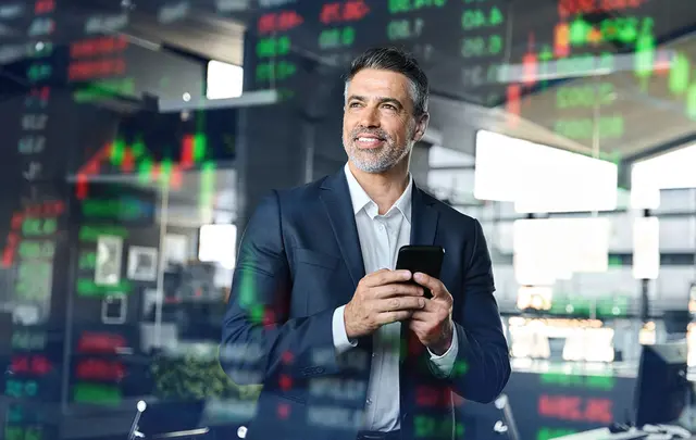 Professional standing in a modern office environment holding a smartphone, with transparent financial market data, stock charts, and trading figures overlaid across the image, illustrating data-driven decision-making, financial analysis, and business intelligence.