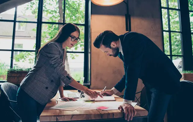 Two professionals collaborate over documents at a wooden meeting table in a modern office with large windows.