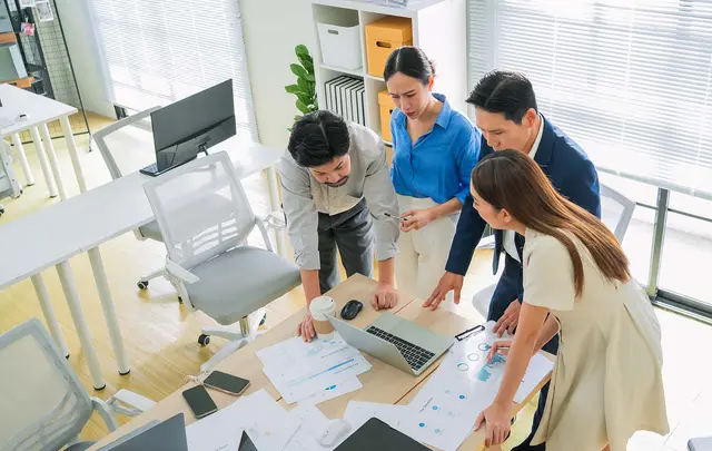 Four professionals gather around a desk in a bright office, reviewing charts and documents alongside a laptop during a collaborative planning and analysis session.