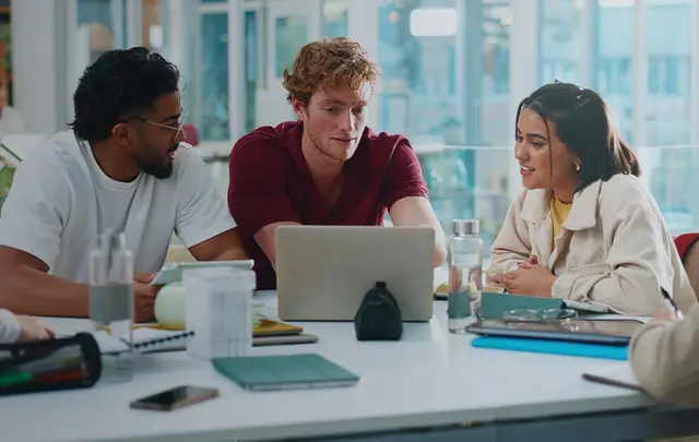 Three professionals collaborating around a laptop in a modern office workspace, reviewing information and discussing projects.