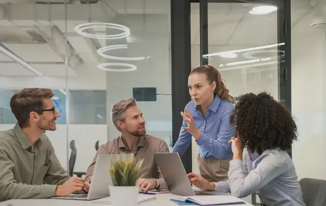 Four professionals collaborate around a conference table in a modern glass-walled office, using laptops while one person stands and presents ideas during a team discussion.